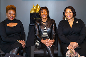 A photo of three black women, smiling at the camera, while seated, at a celebratory event.